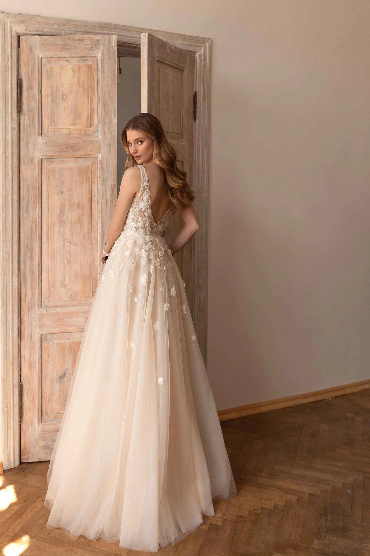 A woman wearing a sleeveless A-line wedding gown with floral applique and a sheer tulle skirt, standing in a room with rustic wooden doors and herringbone wood flooring.