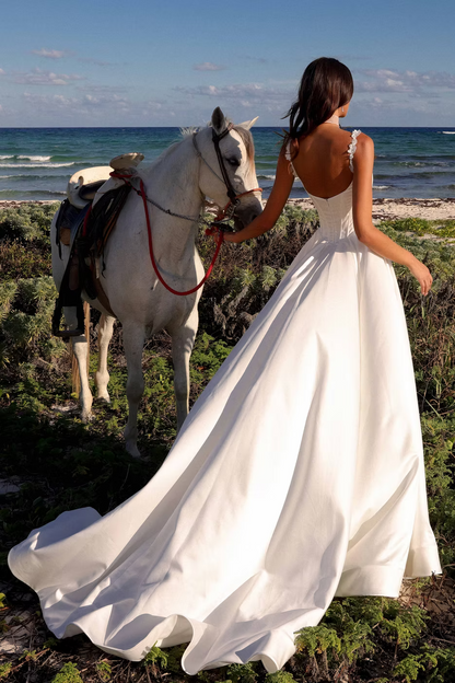 A woman in a flowing white wedding dress stands on a beach with a white horse beside her, photographed from behind to show the back of the dress and the ocean in the background.