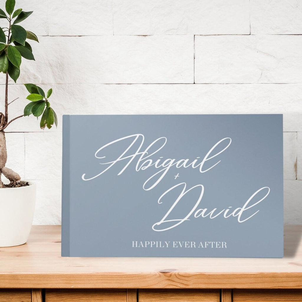 Dusty blue guest book on a table with "Abigail + David, Happily Ever After" written in white calligraphy