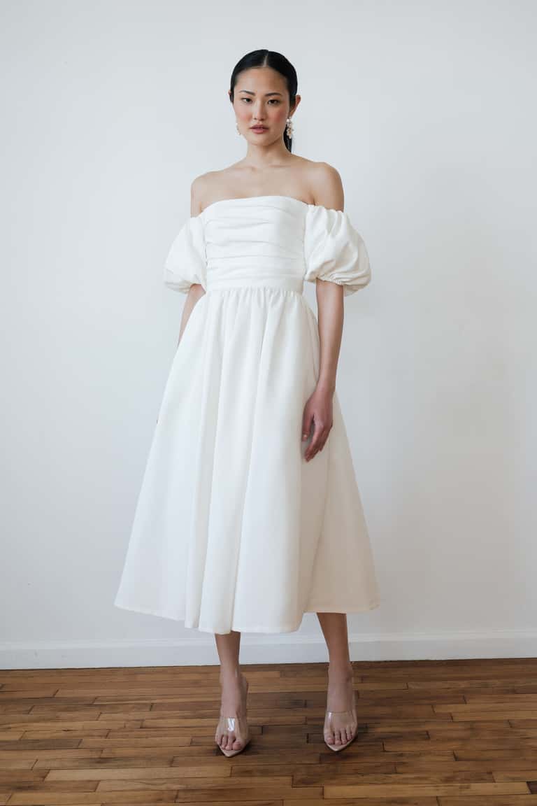 Woman wearing a white off-shoulder dress standing against a plain white wall.
