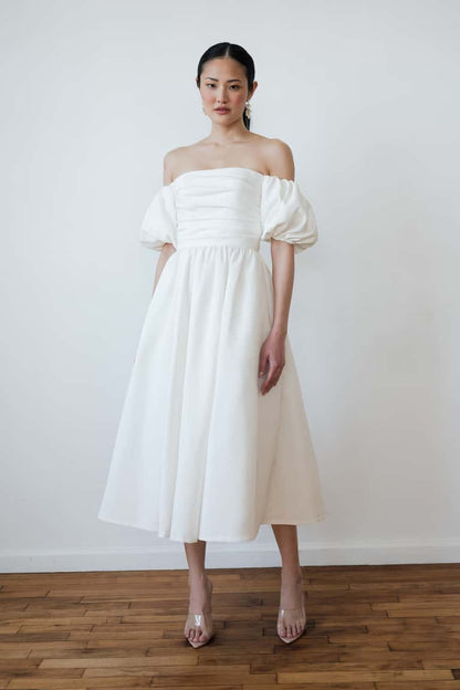 Woman wearing a white off-shoulder dress standing against a plain white wall.