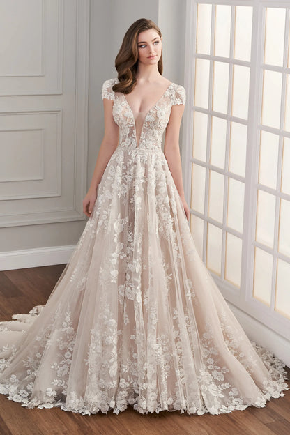 A woman modeling a floor-length A-line wedding gown with a deep V-neckline, cap sleeves, and intricate floral lace applique over a sheer champagne tulle skirt, set in a bright room with a window. Arabella by Martin Thornburg at NKIN