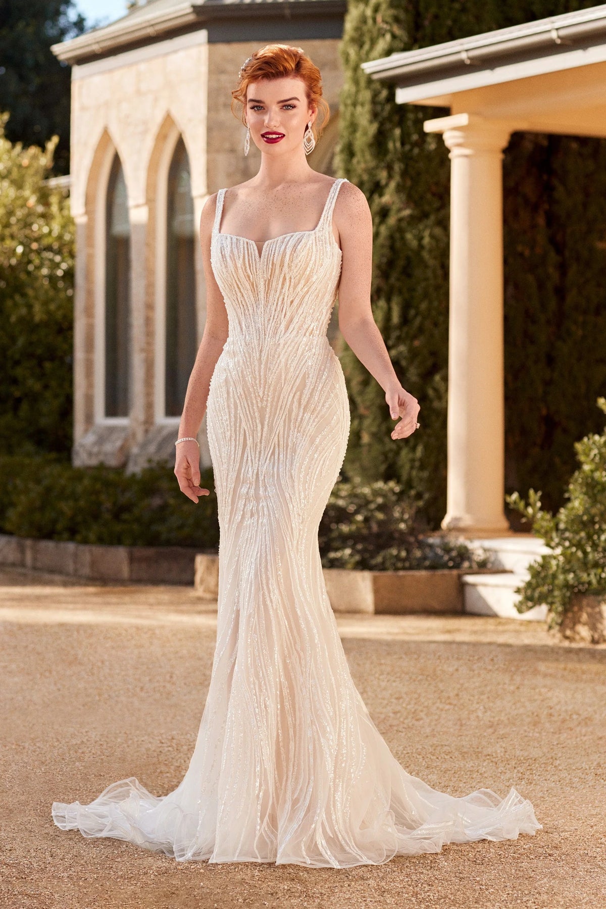 A woman wearing a fitted champagne wedding gown with a train, standing outdoors in front of a stone building with columns and greenery.