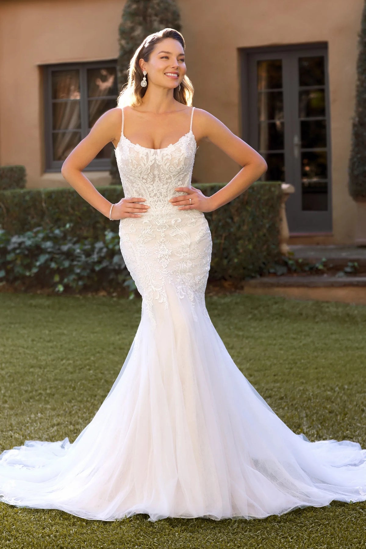 A woman modeling a white lace and tulle mermaid-style wedding gown outdoors on a grassy lawn in front of a beige building with dark-framed windows.