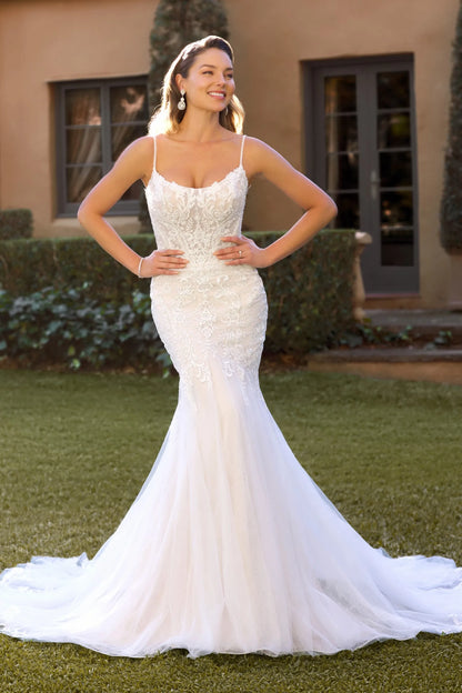 A woman modeling a white lace and tulle mermaid-style wedding gown outdoors on a grassy lawn in front of a beige building with dark-framed windows.