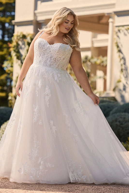 A woman wearing Maya by Sophia Tolli white A-line wedding gown with floral lace applique and a sweetheart neckline, standing outdoors in front of a light-colored building. Available at NKIN bridal boutique in Ottawa