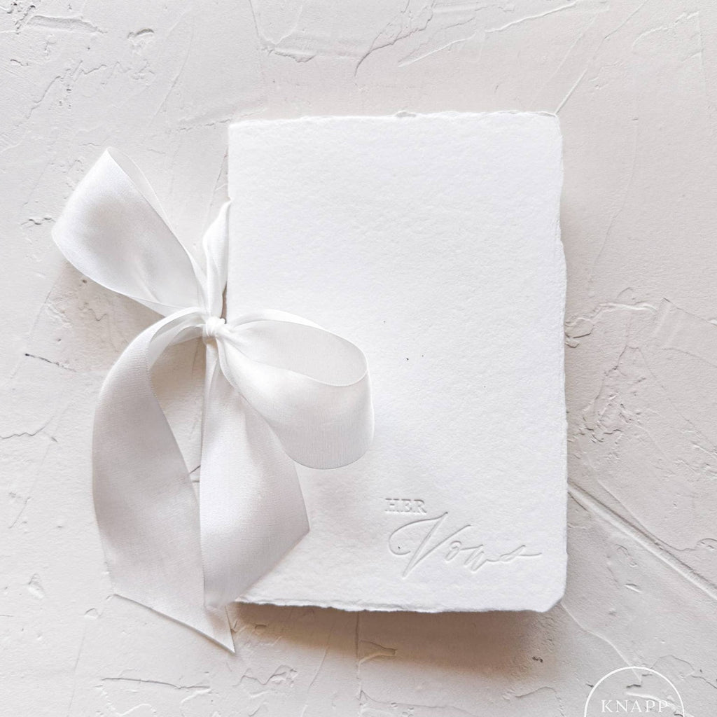 White vow book with white ribbon and "her vows" stamped on bottom right corner