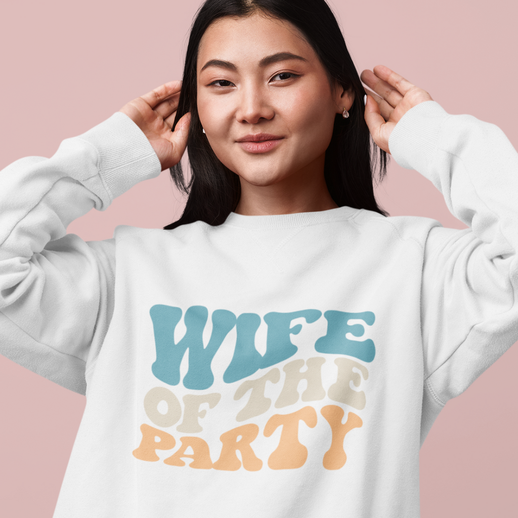 A young asian woman wearing a white crewneck with "wife of the party" written in retro multicolor writing.