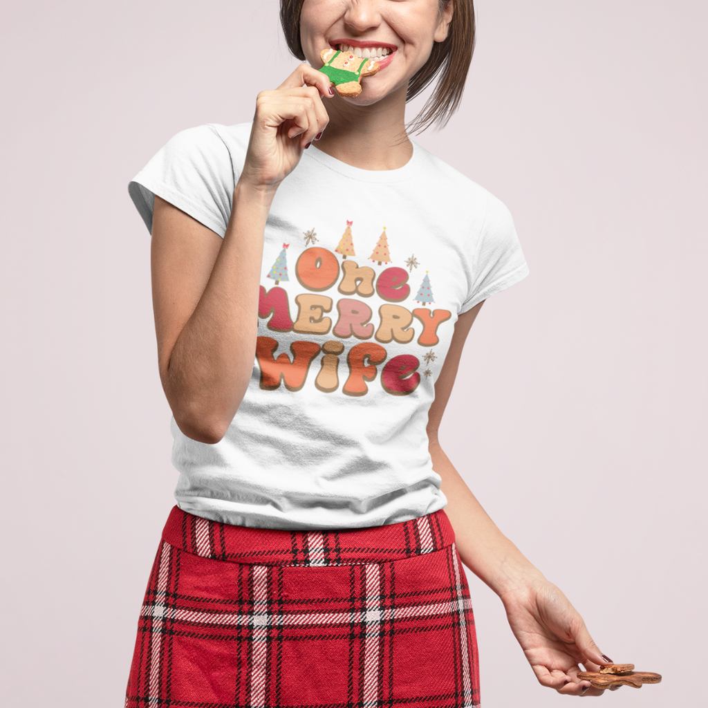 A young white woman wearing a white shirt with "one merry wife" printed on in Christmas design. She is eating a christmas cookie and wearing a red plaid skirt.