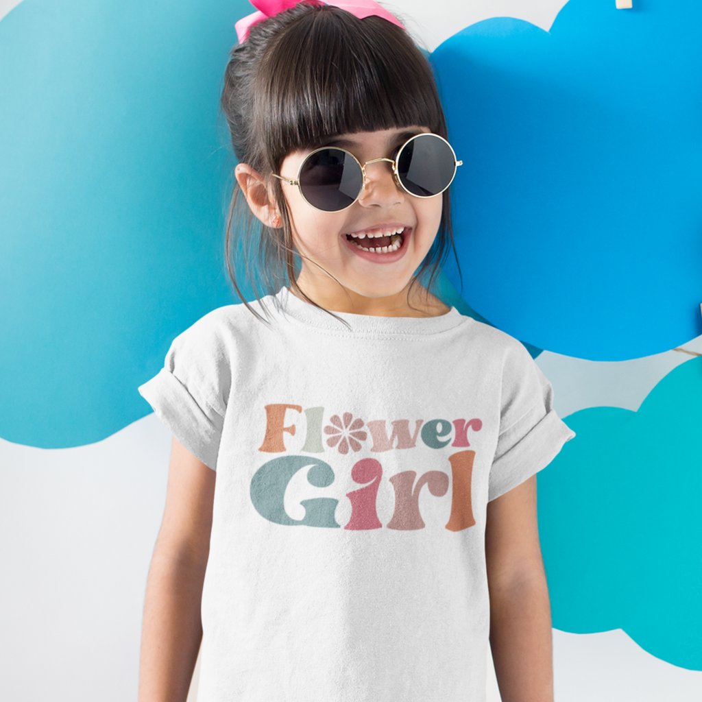 A young girl with sunglasses wearing a white shirt with "flower girl" written in retro writing.