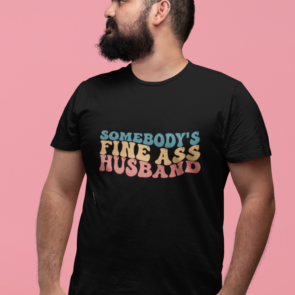 A man wearing a black shirt with "somebody's fine ass husband" written in pastel retro writing.