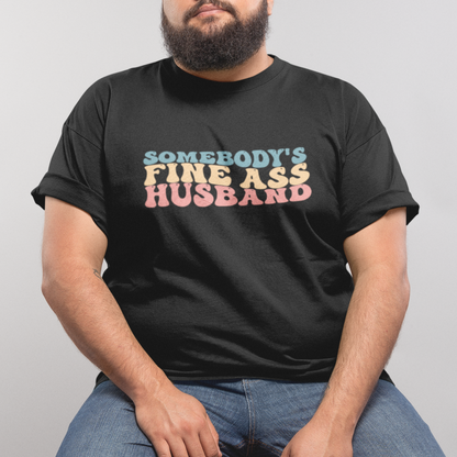 A man wearing a black shirt with "somebody's fine ass husband" written in pastel retro writing.