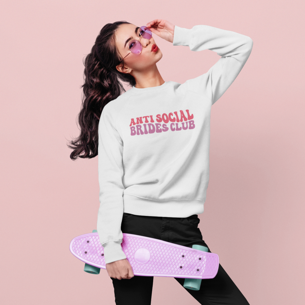 A young woman wearing a white sweatshirt with "anti social brides club" written in pastel retro writing in the centre.