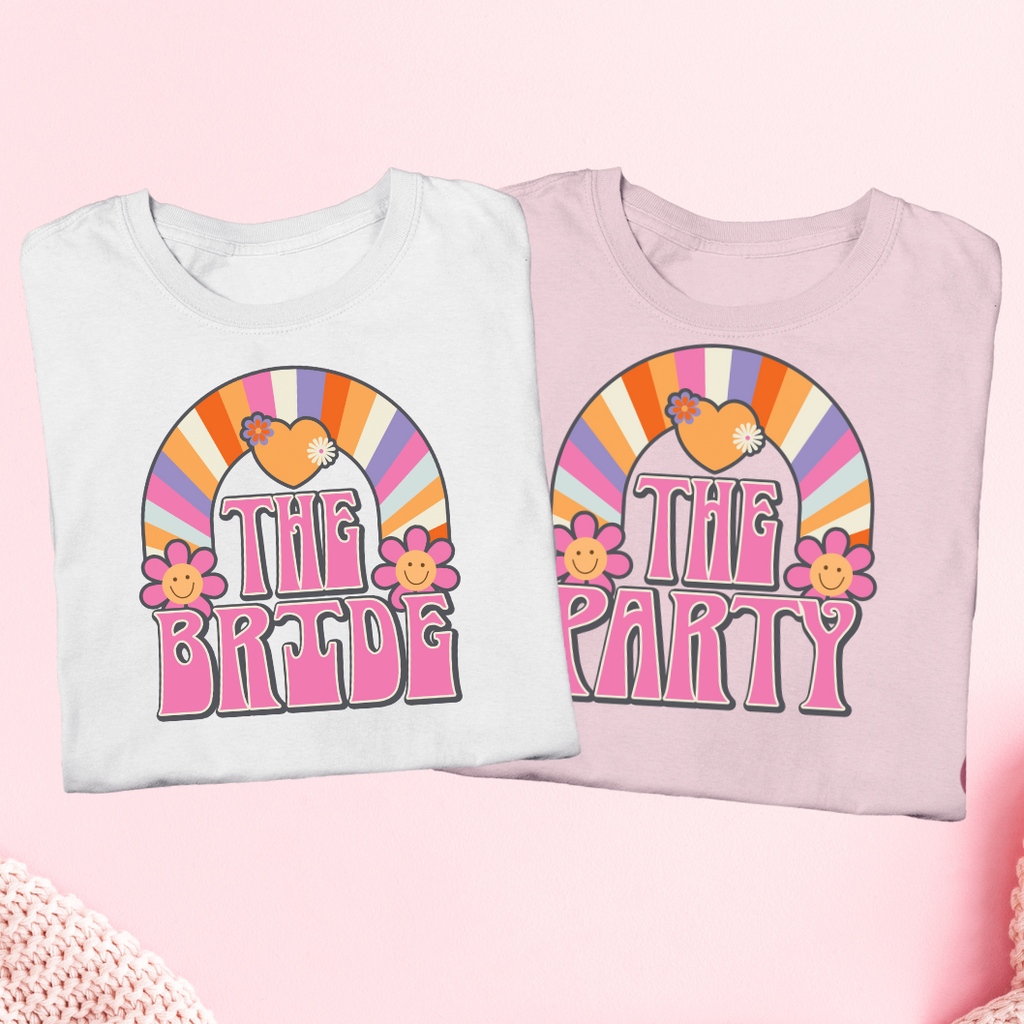 Matching bachelorette party smiley shirts. "The Bride" printed in retro font on a white shirt wiith flowers and a rainbow. "The Party" written in retro font on a pink shirt with flowers and a rainbow.
