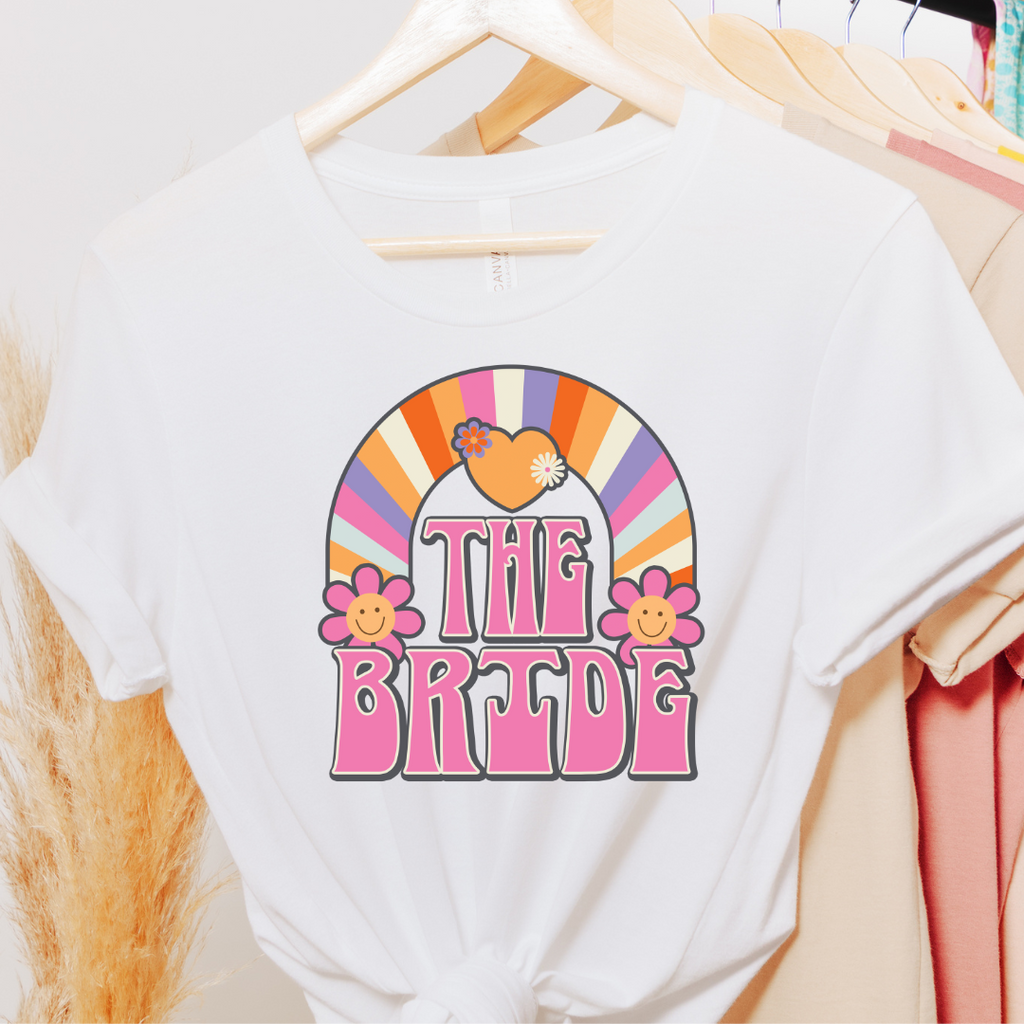 A white T shirt with "The Bride" written in retro pink font with a multicolor rainbow and flowers.