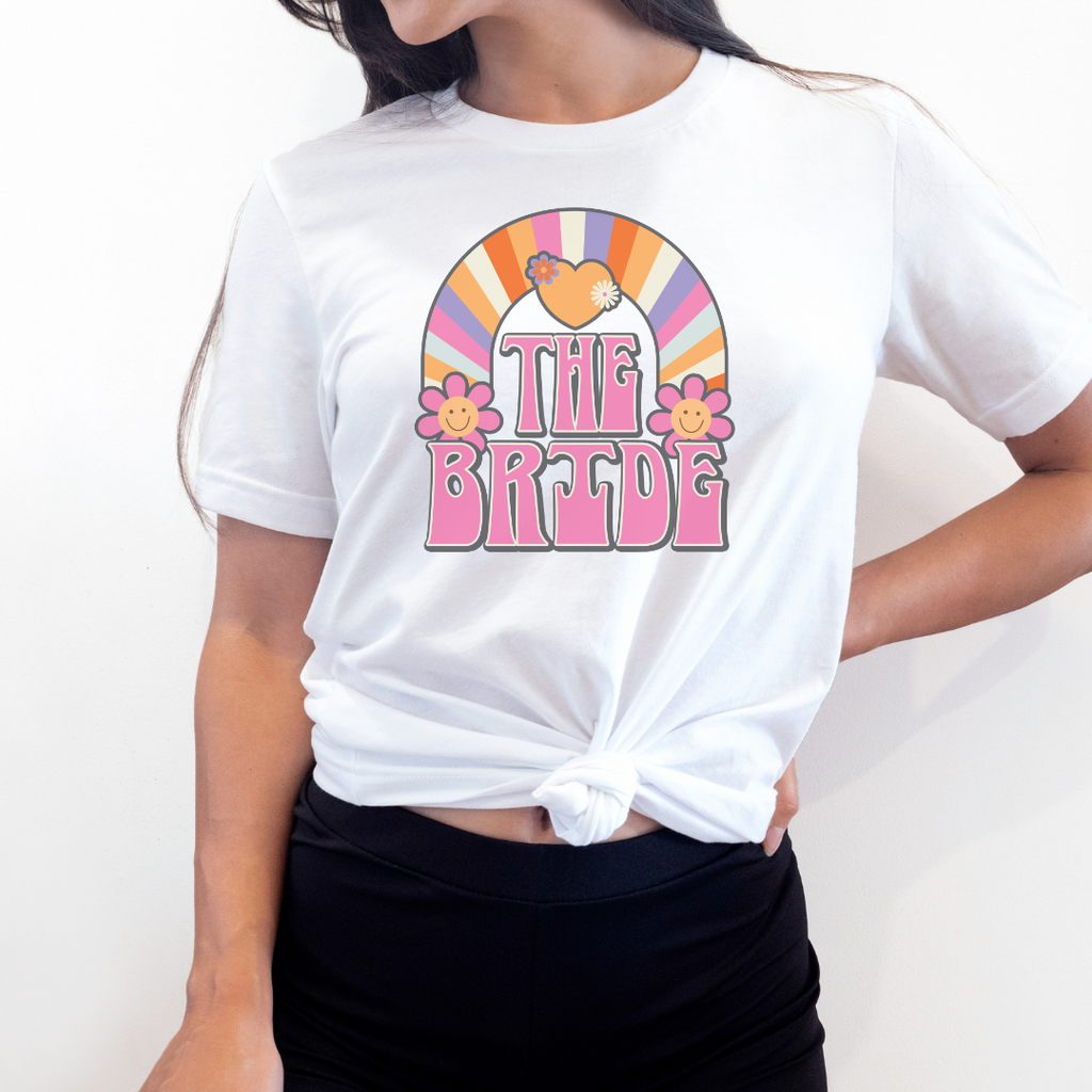 A young woman wearing white T shirt with "The Bride" printed in pink retro font with a multi color rainbow and flowers.