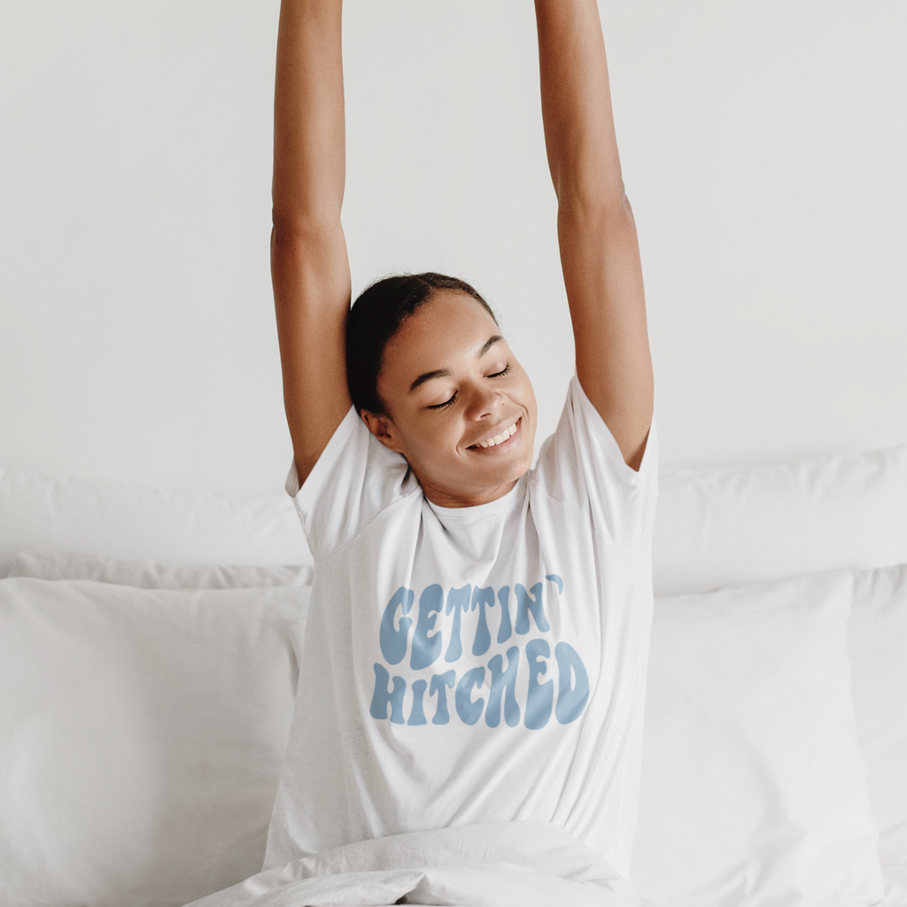 A white shirt with light blue writing saying "Gettin' Hitched" worn by a girl stretching in a white bed. A great inspiration for the morning of the bachelorette party.
