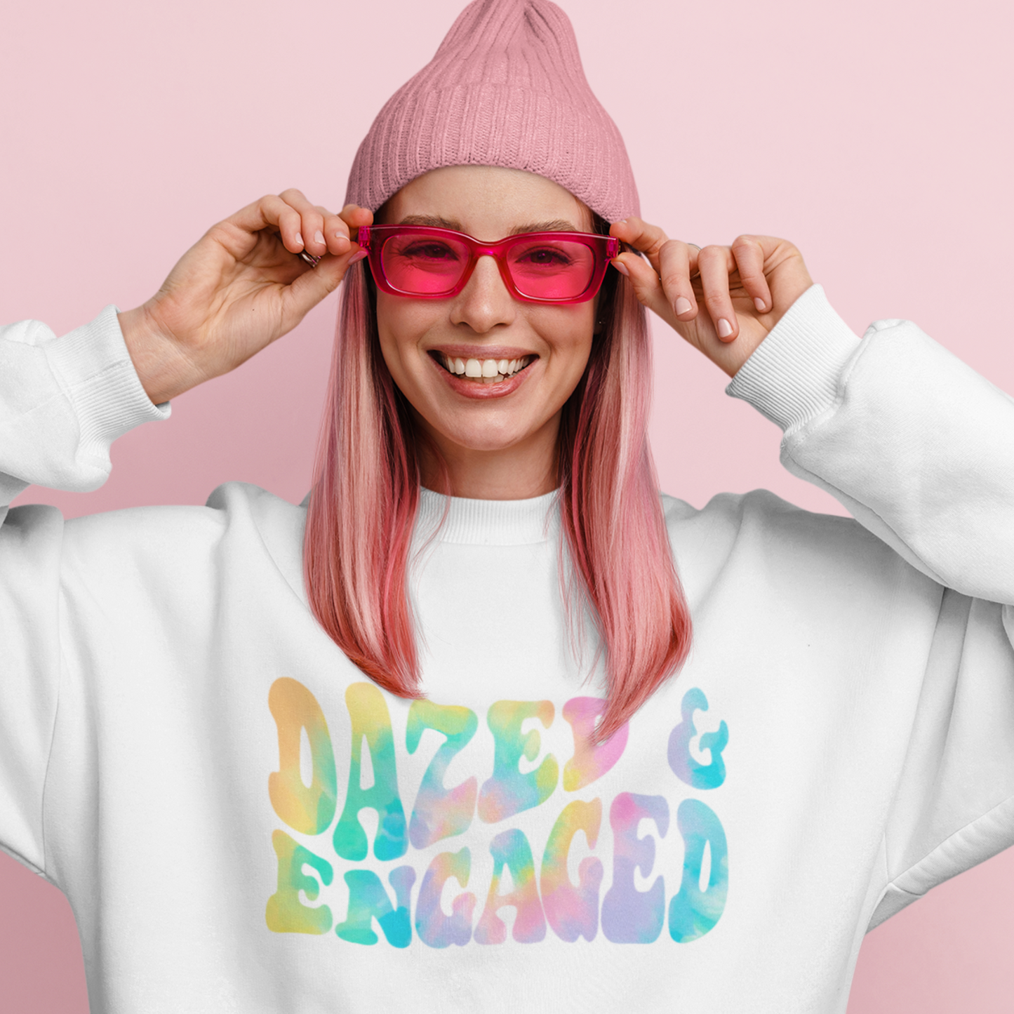 A young woman wearing a hat and red sunglasses with a crewneck with the words "dazed & engaged" written in tie dye. A great gift for the bride, and cozy too!