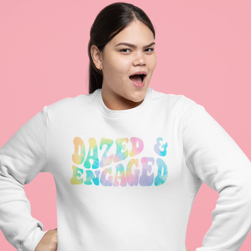 A young happy woman wearing a white crewneck. The crewneck says "dazed & engaged" in a pink, blue and yellow tie dye retro font. What a fun gift for the newlywed. Inclusive and size inclusive.