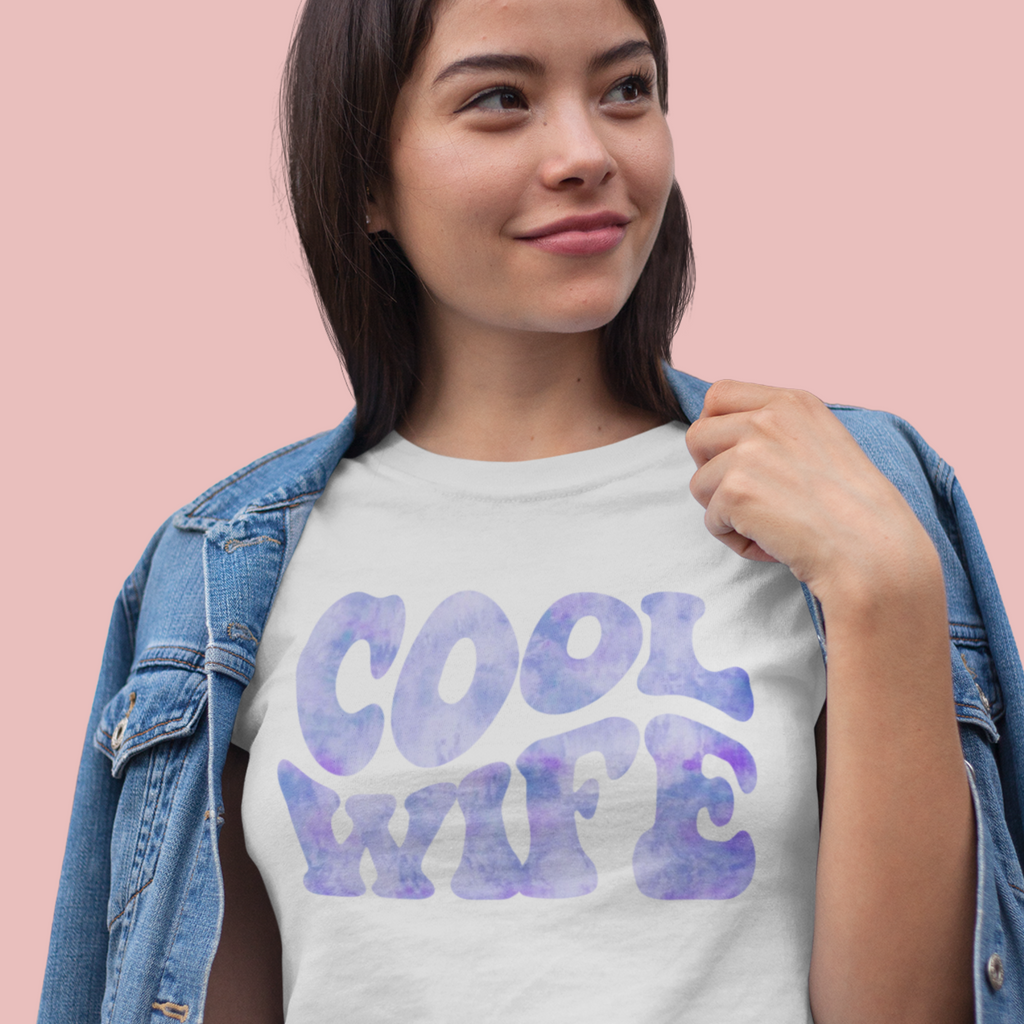 A woman wearing a jean jacket and white T shirt with "cool wife" written in tie dye blue. In front of a pink background. The perfect engagement gift for the bride!