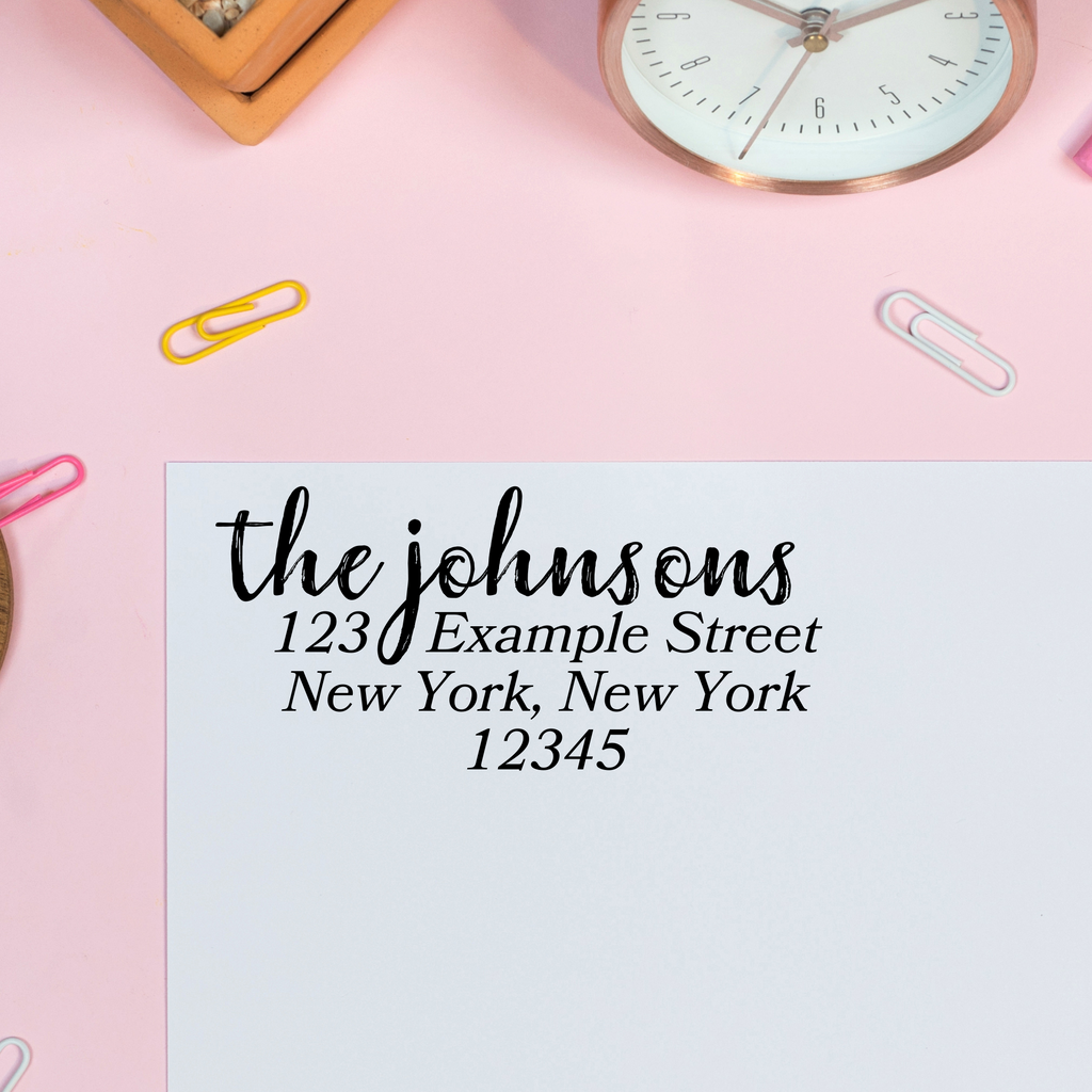 A stamp on an envelope - "the johnsons" written in calligraphy, "123 Example Street, New York New York, 12345" A pink background with stationary supplies surrounds. 