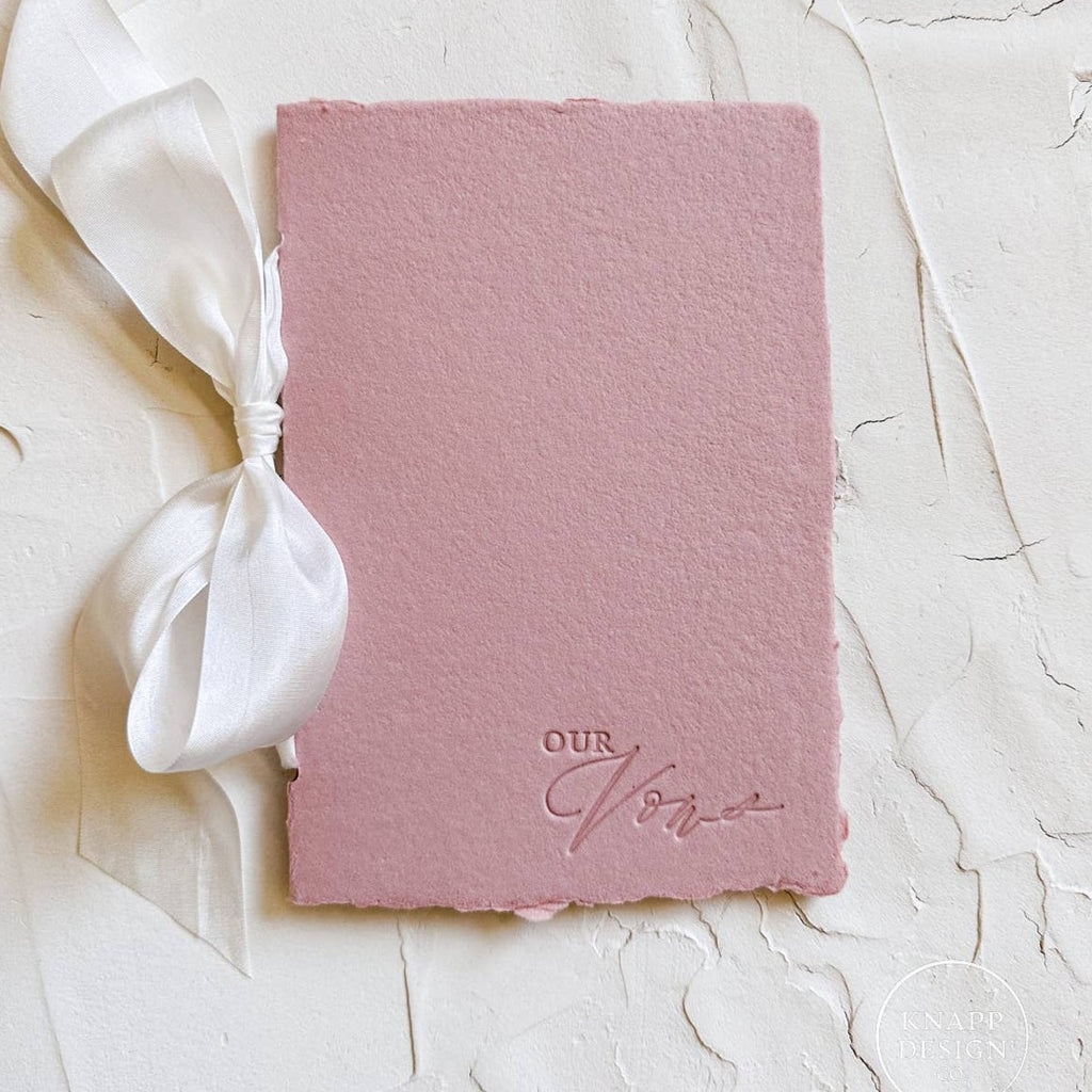 Pink vow book with white ribbon and "our vows" pressed in the bottom right corner