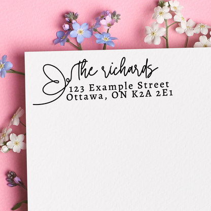 A black self inking stamp on a white nevelope. "The richards" written in cursive with a butterfly silhouette. The rest of the address is written in block writing. Sat on a floral pink background.