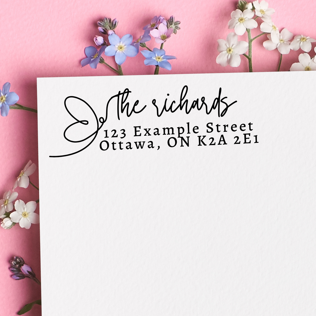 A black self inking stamp on a white nevelope. "The richards" written in cursive with a butterfly silhouette. The rest of the address is written in block writing. Sat on a floral pink background.