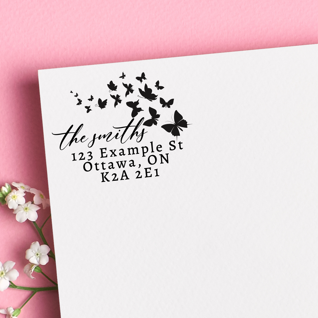 A black self inking stamp on a white envelope. Butterfly silhouettes surround the writing. "The smiths" written in calligraphy and the rest in block script. The envelope is on a floral pink background.  A super unique housewarming gift.