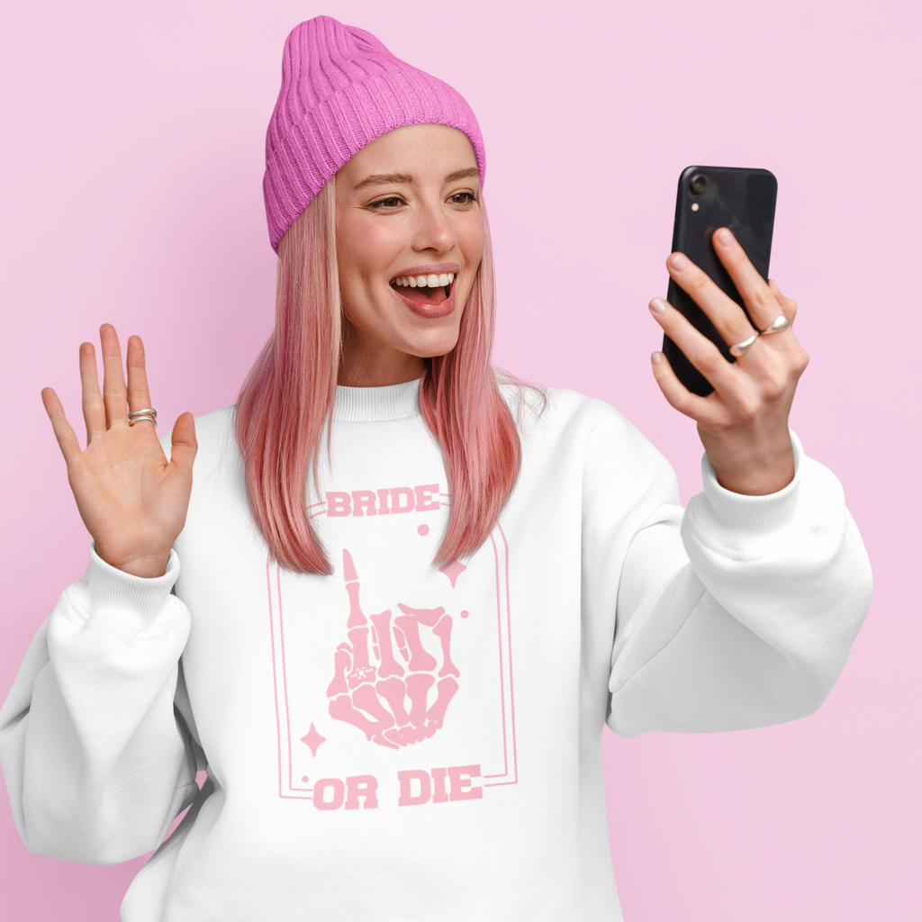 A young woman with pink hair and a pink beanie on the phone. She is wearing a white crewneck sweater with "bride or die" printed in pink with a skull hand on it. A unique bride to be gift.