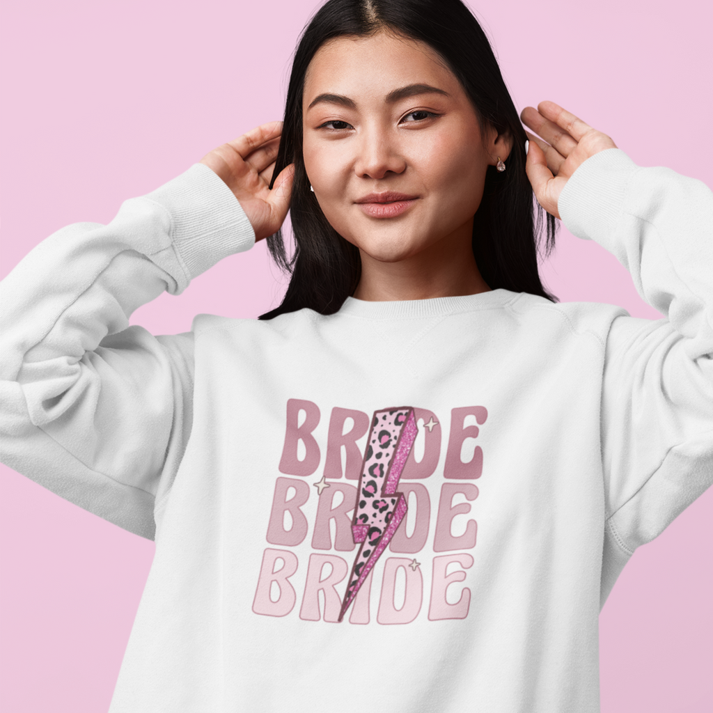 A young woman wearing a white sweatshirt. "Bride" is repeated three times in pink with a leopard lightning bolt on top. A super cool and retro bride to be gift.