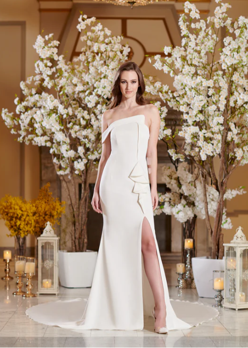 Woman wearing a wedding dress with an asymmetrical neckline, slit up the front leg and ruffled embellishments